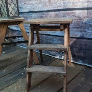 A miniature handcrafted raw wood step ladder styled like a vintage painter's stool with black metal hinges sitting on a rustic workshop bench.